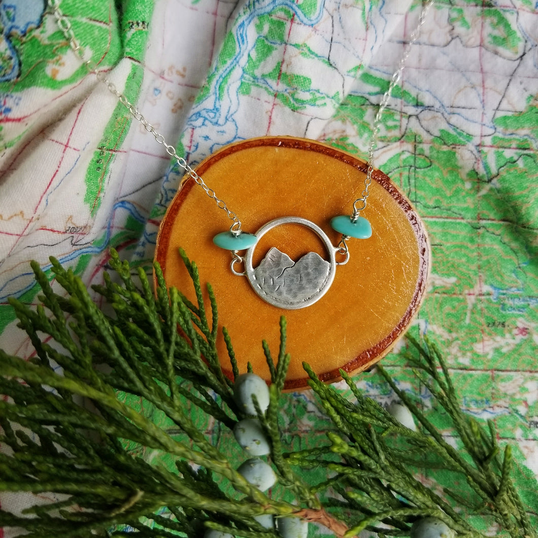 silver mountain necklace with turquoise beads resting on a wood slice on top of a map with a juniper sprig
