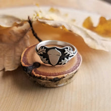 An acorn signet ring front view, sitting atop a tiny wood slice with golden oak leaves in the background