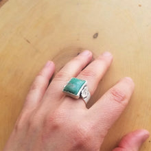 A hand wearing an aspen ring with a blue green stone on the middle finger, resting atop a wooden background