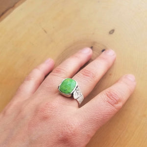 A hand wearing an aspen ring with a bright green stone on the middle finger, resting atop a wooden background