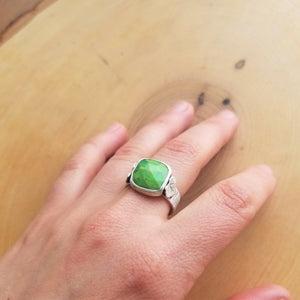 A hand wearing an aspen ring with a bright green stone on the middle finger, resting atop a wooden background