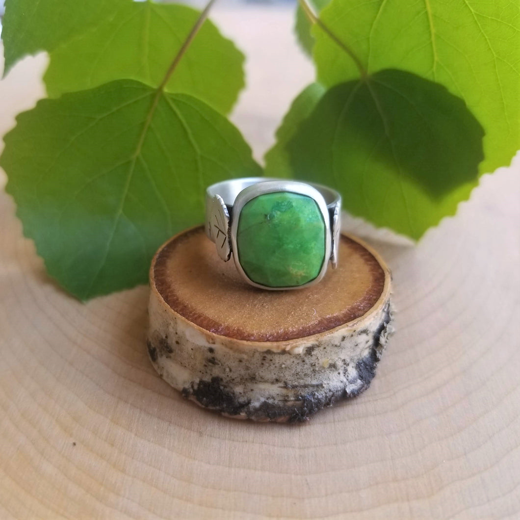 An aspen ring with a bright green stone sits atop a tiny wood slice, with green aspen leaves in the background