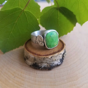 An aspen ring with a bright green stone, side view, sits atop a tiny wood slice, with green aspen leaves in the background