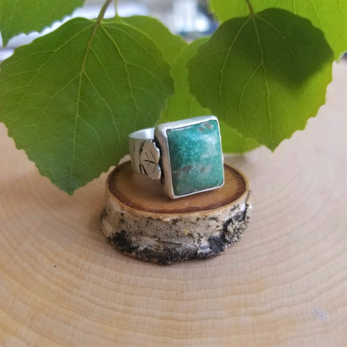 An aspen ring with a blue green stone, side view, sits atop a tiny wood slice, with green aspen leaves in the background