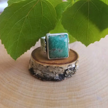 An aspen ring with a blue green stone, front view, sits atop a tiny wood slice, with green aspen leaves in the background