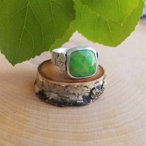 An aspen ring with a bright green stone, side view, sits atop a tiny wood slice, with green aspen leaves in the background