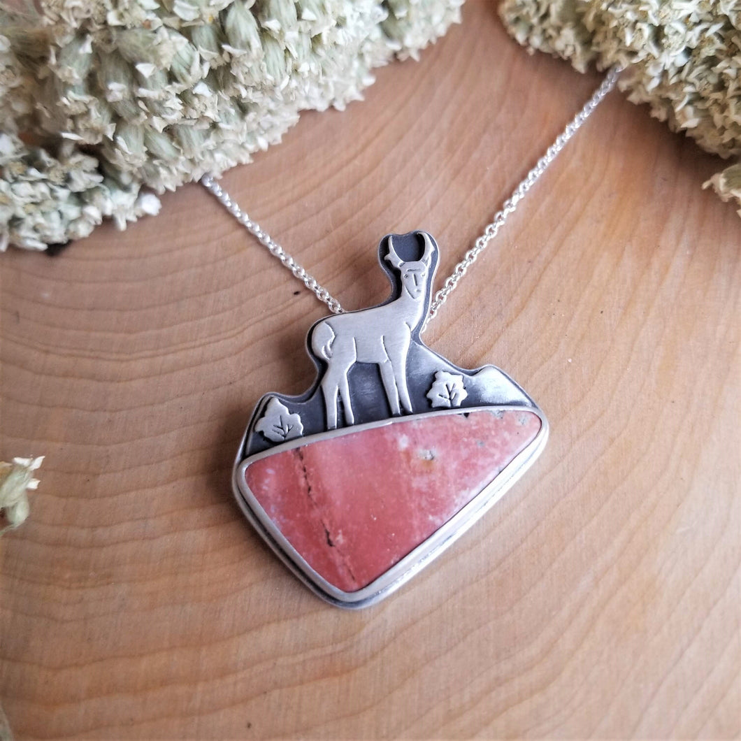 A necklace sitting on a wooden background with yarrow flowers. The necklace has an antelope standing in front of a mountain background made of silver, with a pink jasper stone underneath. 