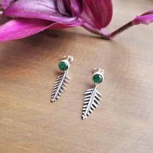 Stud earrings with green gemstones and tiny dangling ferns, sits on a wooden background with purple leaves.