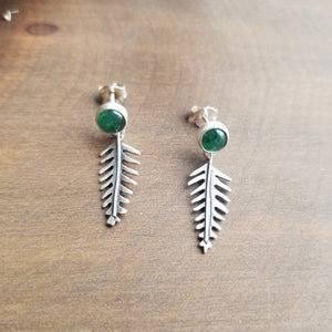 Stud earrings with green gemstones and tiny dangling ferns, sits on a dark wooden background.