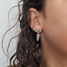 A person's ear with curly brown hair and multiple piercings, wearing a stud earring with a green gemstone and tiny dangling fern. 