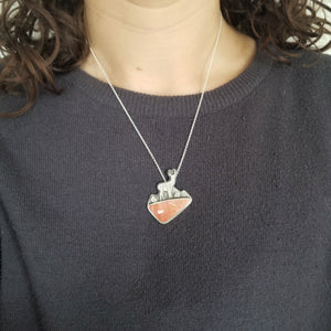 A necklace with an antelope standing in front of a mountain background made of silver, with a pink jasper stone underneath. The necklace is worn around a person's neck, over a dark gray shirt.