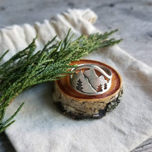 silver mountain ring with pine tree silhouettes resting on a wood slice and linen bag with a juniper sprig